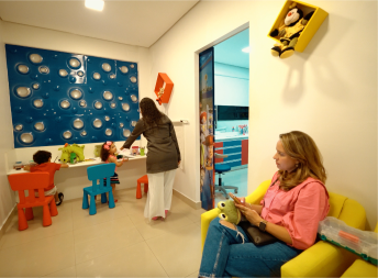 Doutora Adrielly chamando a paciete criança para entrar no consultório, enquano outros pacientes e seus pais aguardam na sala de espera infantil