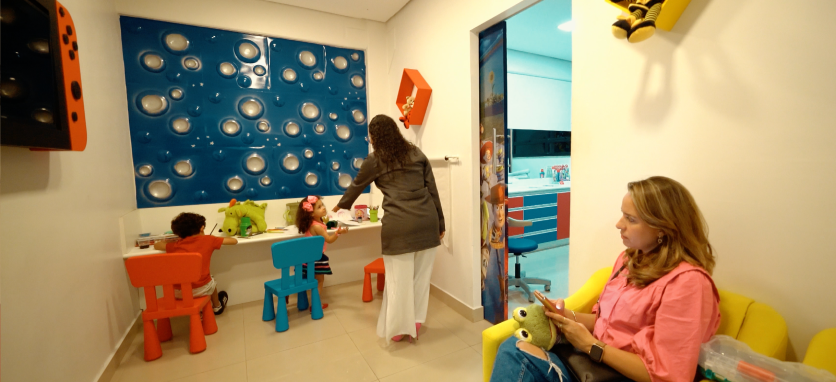 Doutora Adrielly chamando a paciete criança para entrar no consultório, enquano outros pacientes e seus pais aguardam na sala de espera infantil