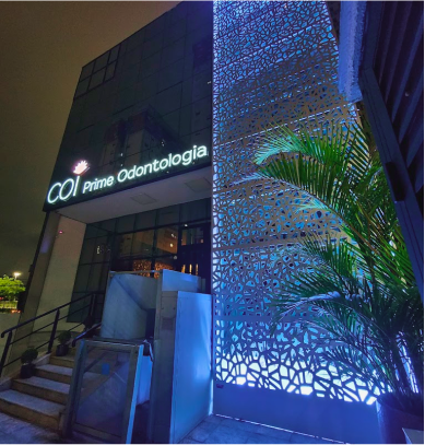 A faixada da clínica a noite com uma luz azul e o letreiro onde está escrito COI Prime Odontologia aceso com uma luz branca