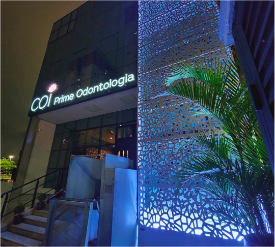 A faixada da clínica a noite com uma luz azul e o letreiro onde está escrito COI Prime Odontologia aceso com uma luz branca